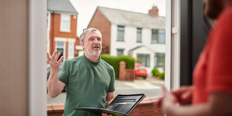 A man holding a folder talking to a householder at an open front door on a residential street
