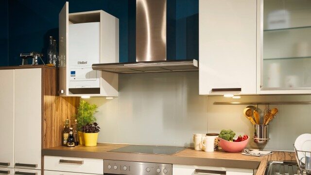 A white Vaillant wall-mounted boiler installed in an open kitchen cupboard above a worktop with an induction hob and extractor hood