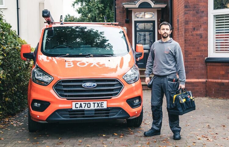 A Boxt engineer holding a tool bag standing next to an orange Boxt-branded Ford Transit van outside a brick house