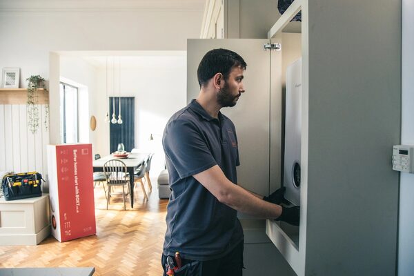 An engineer in a navy polo shirt installing a wall-mounted boiler inside a kitchen cupboard, with a red Boxt delivery box in the background