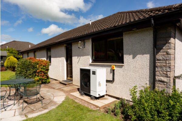 An air source heat pump unit installed on the exterior wall of a rendered bungalow with a patio and garden