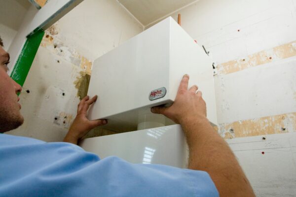 An installer fitting a white Alpha boiler casing onto a wall-mounted boiler in an unfinished room