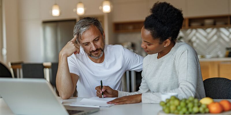 A couple at a kitchen table reviewing paperwork together with a laptop open in front of them