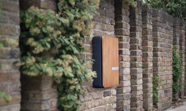 An Andersen EV wooden-cased electric car charge point mounted on an outdoor brick wall beside climbing plants