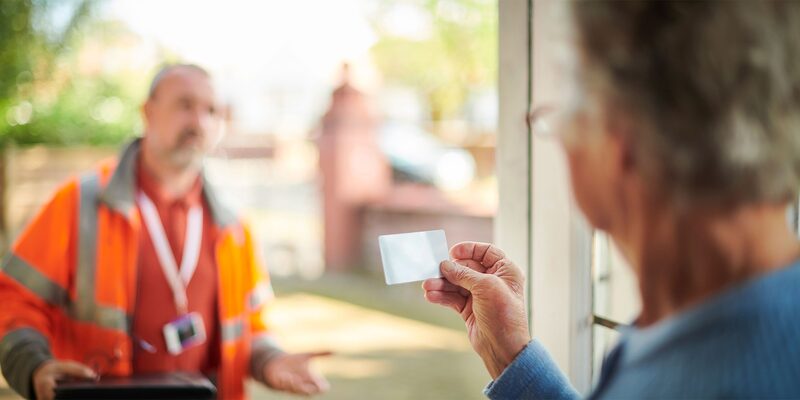A householder at their front door inspecting an ID card held up against a workman in a high-visibility orange jacket