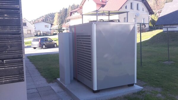 A grey outdoor air-source heat pump unit installed on a concrete slab beside a house, with a car and houses in the background