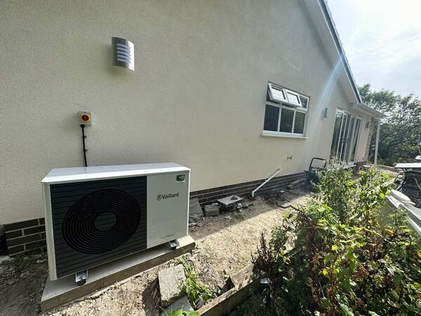 A Vaillant air source heat pump outdoor unit installed on a concrete base against the white rendered wall of a house, with shrubs and rubble in the foreground.