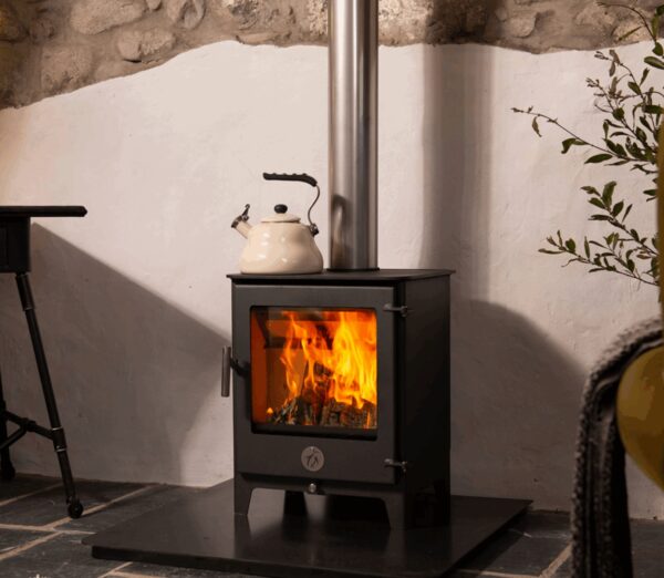 A black wood-burning stove with flames visible through the glass door, a kettle on top, installed in a room with a stone wall