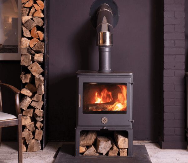 A black wood-burning stove with flames burning inside, installed in a room with a log store and flue pipe