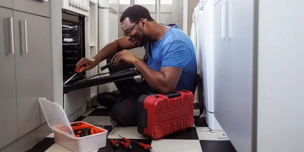 A tradesman kneeling in a kitchen repairing an oven door with a red toolbox and screwdrivers beside him