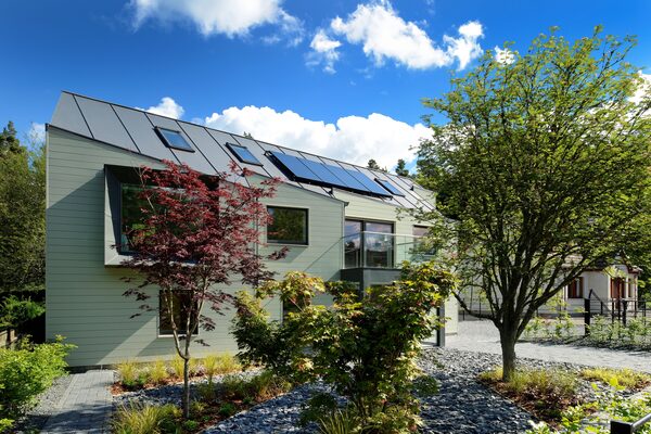 A modern energy-efficient house with solar panels on its metal roof, surrounded by trees and garden