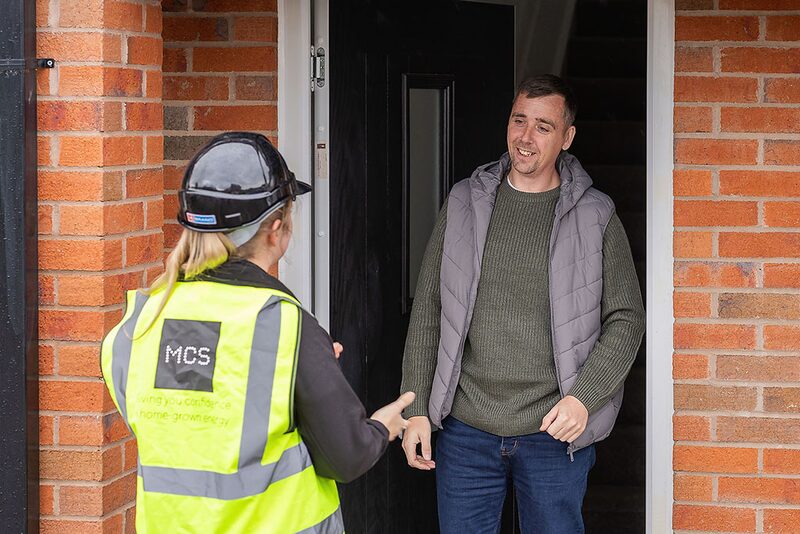 An installer in an MCS-branded hi-vis vest and hard hat talking with a homeowner at a brick house doorway