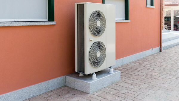 An air source heat pump unit mounted on a concrete plinth against the terracotta exterior wall of a house