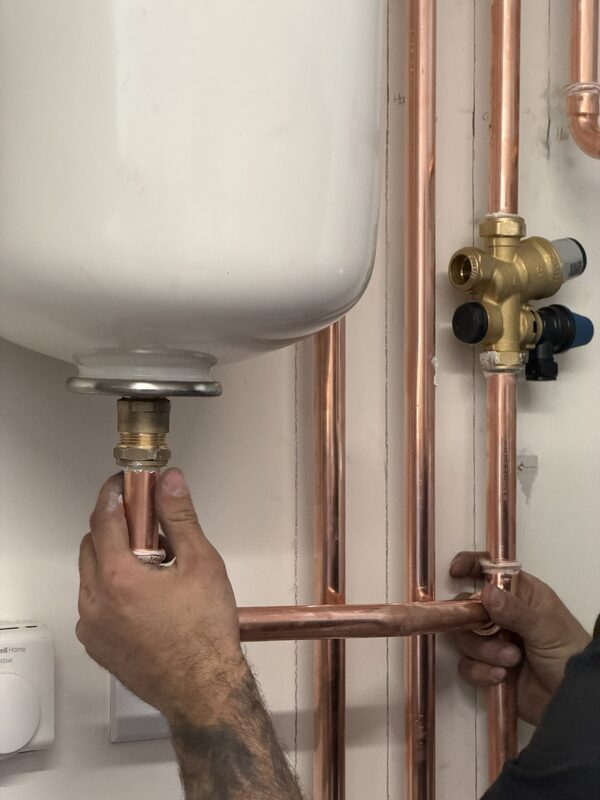 A plumber's hands connecting copper pipework to a white hot water cylinder with brass valves during a heat pump installation