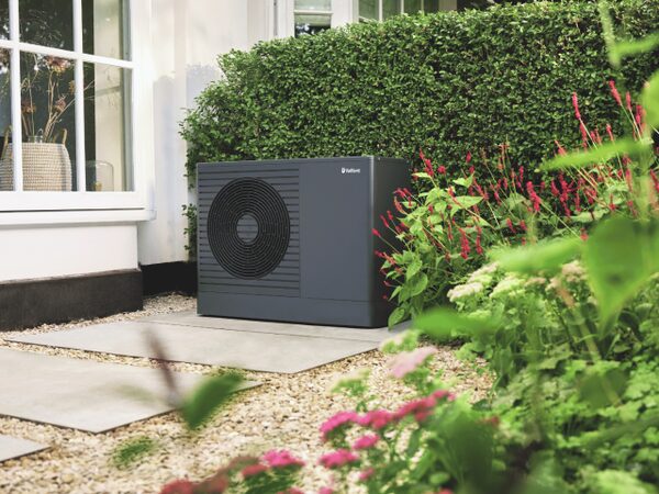 A Vaillant aroTHERM pro air source heat pump installed outside a house beside a hedge and flower bed.