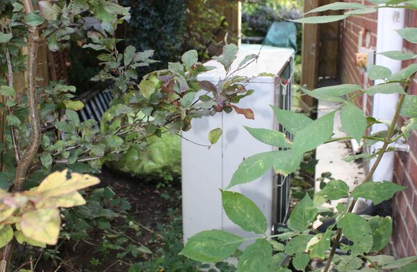 An outdoor air source heat pump unit partly hidden by overgrown plants and foliage beside a brick house wall