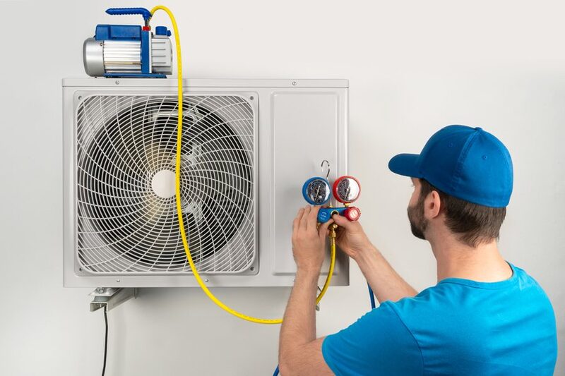 A technician in a blue cap using manifold gauges and a vacuum pump to commission a wall-mounted air source heat pump unit