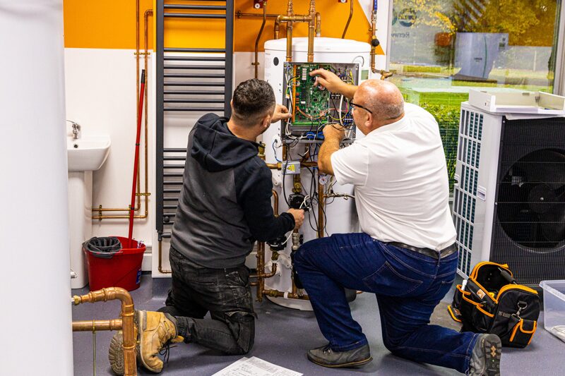 Two engineers working on the wiring of a heat pump cylinder installation in a training room, with an air source heat pump unit beside them