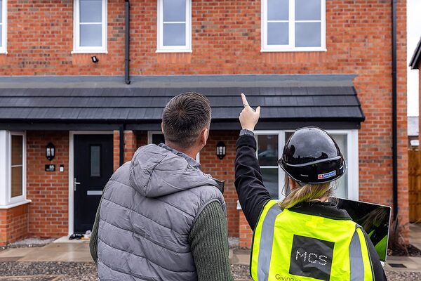 An MCS assessor in a hi-vis vest and hard hat points at the roof of a brick house while talking to a homeowner