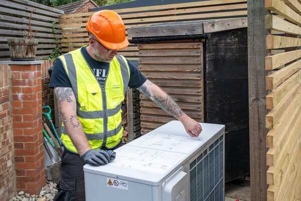 An engineer in a hi-vis vest and orange hard hat installing an air source heat pump unit in a garden
