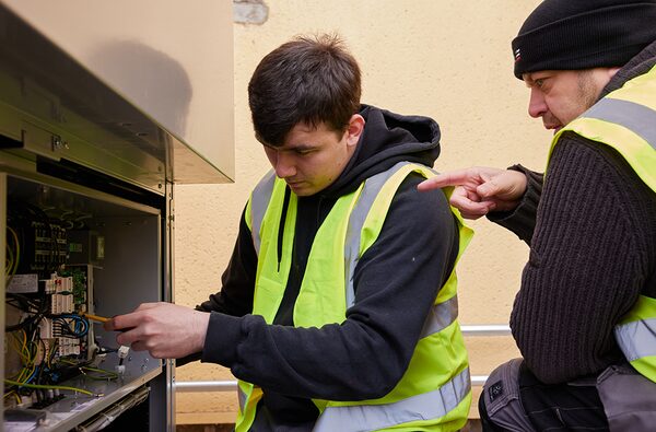 Two engineers in high-visibility vests working on the wiring inside a heat pump unit outdoors