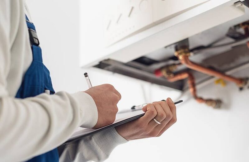 An engineer in blue overalls writing on a clipboard while servicing a wall-mounted boiler with copper pipework