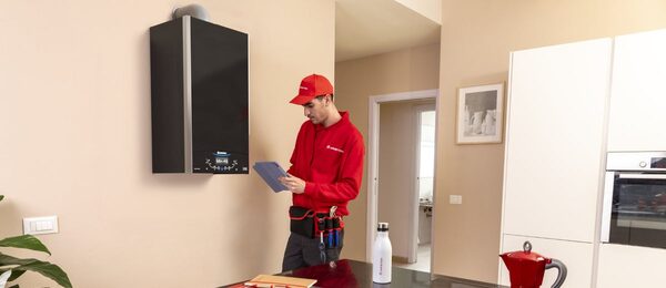 An Ariston engineer in red uniform inspecting a wall-mounted black boiler in a home kitchen