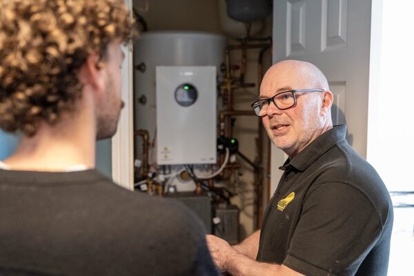 An installer in a dark polo shirt talking to a homeowner beside a wall-mounted heat pump cylinder and hot water tank in an airing cupboard