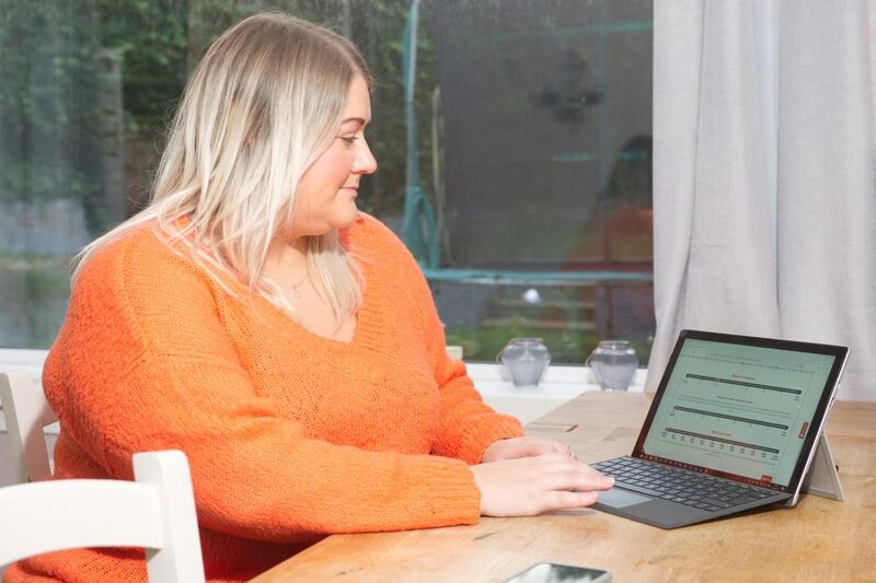 A woman in an orange jumper using a laptop at a kitchen table, viewing what appears to be an online portal form