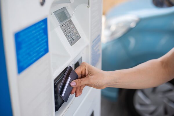A hand inserting a payment card into a card reader on a charging or payment terminal, with a blue car blurred in the background
