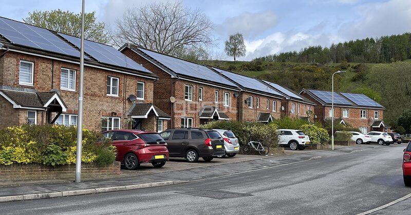 A row of brick terraced houses with rooftop solar panels, parked cars along the street