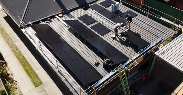 Installers fitting black solar panels on a metal roof surrounded by scaffolding, seen from above