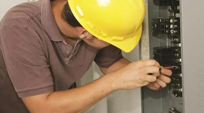 An electrician in a yellow hard hat working on a consumer unit with a screwdriver