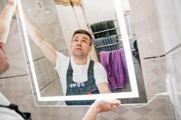 An electrician in overalls fitting an illuminated LED bathroom mirror on a tiled bathroom wall