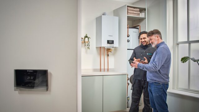 An installer and a homeowner looking at a phone next to a wall-mounted Vaillant boiler and cylinder in a utility room, with a Vaillant thermostat on the wall