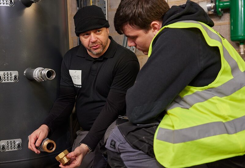 Two engineers inspecting fittings on a hot water cylinder in a plant room, one wearing an MCS-branded shirt