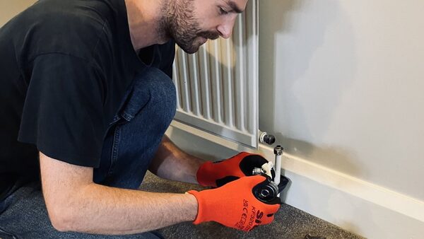 An installer in orange gloves fitting pipework beneath a white radiator in a home