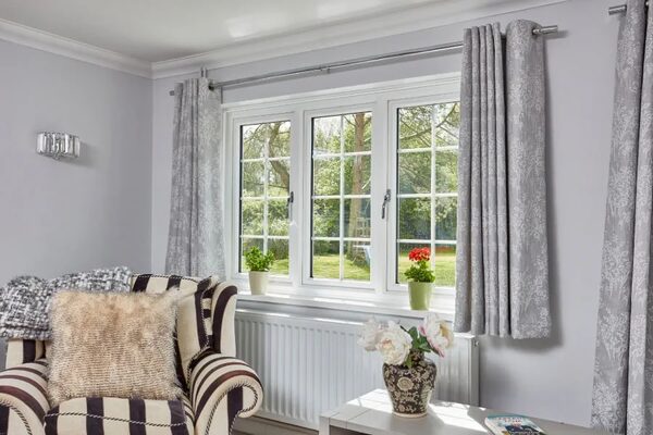 A white casement window with glazing bars installed in a grey living room with curtains, an armchair and a radiator below