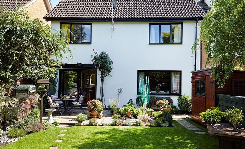 A white two-storey UK house with black-framed windows and a patio door, seen from a garden with lawn, patio furniture and a wooden shed