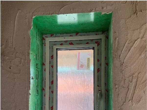 A newly installed uPVC window with protective film still on the frame, set in a reveal lined with green airtightness membrane and unfinished plaster
