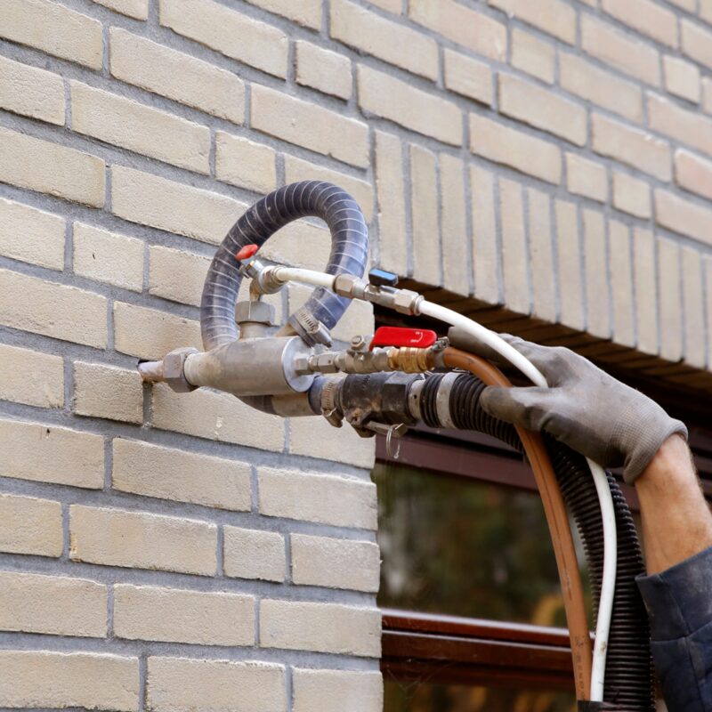 A gloved worker injecting cavity wall insulation into a brick wall through a nozzle and hoses