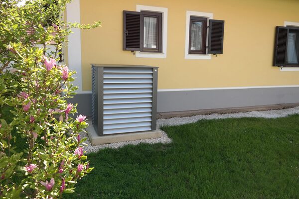 An outdoor heat pump unit with a grey louvred cover installed on a gravel base beside the yellow exterior wall of a house