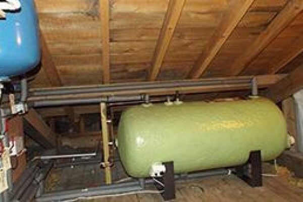 A large green horizontal water storage cylinder installed in a loft with pipework and a blue expansion vessel