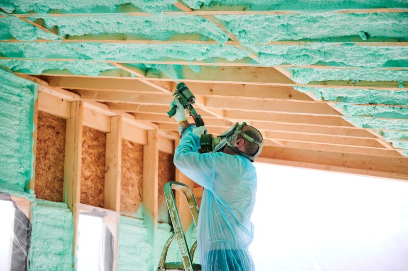 A worker in protective suit and respirator on a ladder spraying green foam insulation between roof joists in an unfinished timber-framed roof