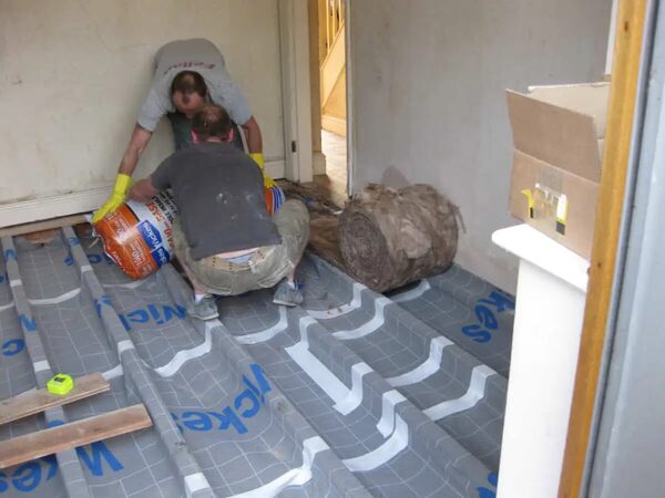 Two workers installing underfloor insulation between floor joists, with a roll of mineral wool and reflective membrane