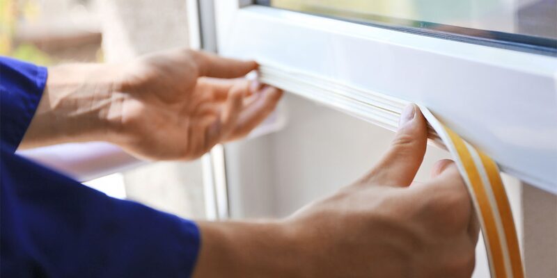 A person's hands applying self-adhesive draught-excluding foam strip to a white window frame
