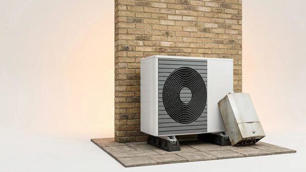 A white air source heat pump unit installed outside against a brick wall, with an old rusty boiler tipped over beside it