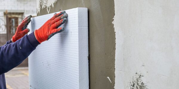 A worker in orange gloves fitting a white foam insulation board to an exterior wall covered in adhesive