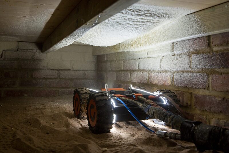 A Q-Bot robotic underfloor insulation robot working beneath floorboards in a crawl space, spraying insulation onto the underside of floorboards