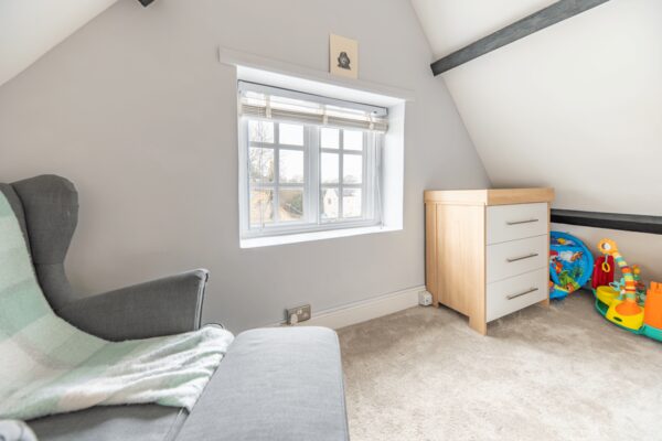 A white horizontal sliding secondary glazing unit installed on a small window in an attic bedroom with a grey armchair and a changing table